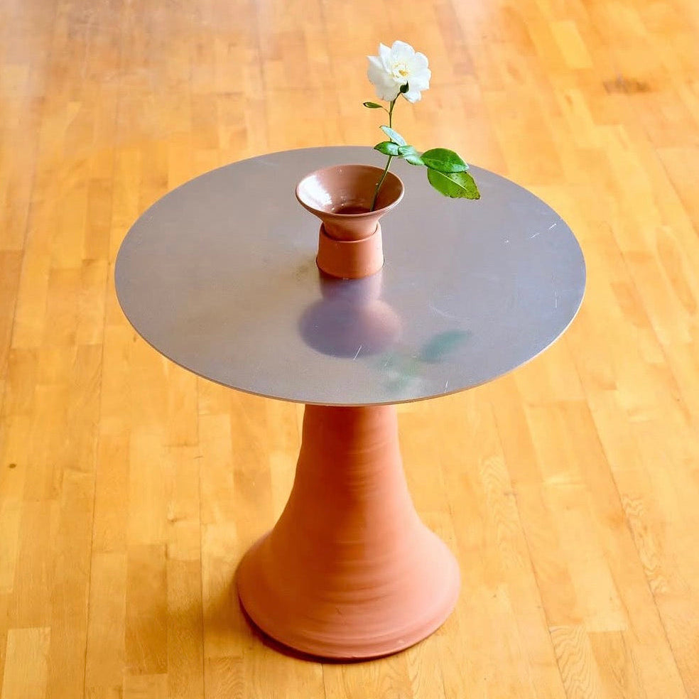 Small round table with a terracotta pedestal base and aluminium top on a wooden floor, featuring a vase with a white flower.